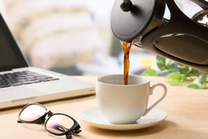 パソコンの前にあるカップにコーヒーを入れている画像。